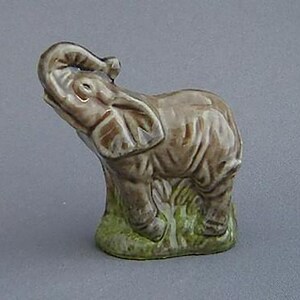 May include: A small ceramic figurine of an elephant with its trunk raised in the air. The elephant is brown and grey with green grass at its feet.