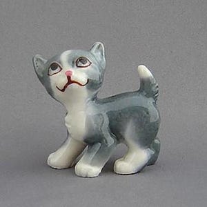 May include: A ceramic kitten figurine. The kitten is depicted in a playful stance, with a grey and white coat, a pink nose, and a smiling expression. The figurine is standing on all fours with its tail raised.