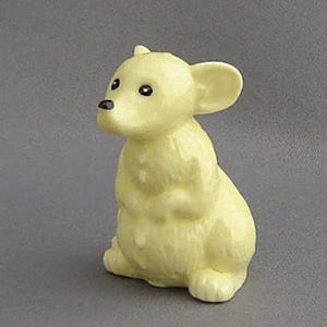 May include: A small, yellow ceramic figurine of a mouse sitting up with its paws crossed. The mouse has black eyes and a black nose.