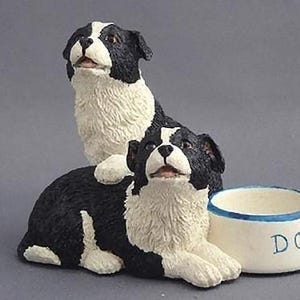 May include: Two black and white border collie dog figurines sitting next to a white ceramic dog bowl with blue trim. The bowl says "DOG" on the front.