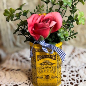 Könnte beinhalten: Eine gelbe, sechseckige Metalldose mit der Aufschrift Franklin's Famous Java Coffee in schwarzer Schrift und Grafik. Ein blau-weiß kariertes Band ist oben um die Dose gebunden. Sie enthält eine große rosa Rose und grüne Blattstiele.