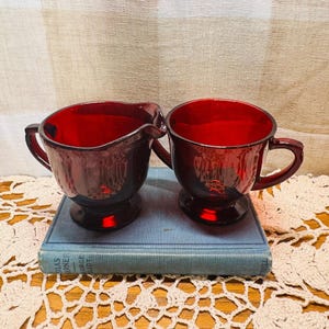 May include: A pair of deep red glass vessels, consisting of a creamer with a pouring spout and a matching mug, both featuring a pedestal base and a handle. The vessels are positioned on top of a blue hardcover book with visible text on the spine.