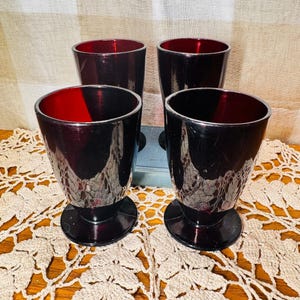 Puede incluir: Un juego de cuatro vasos de vidrio de color rojo intenso con una silueta cónica y una base circular. Cada vaso presenta un acabado translúcido de color rubí oscuro que refleja la luz, creando una apariencia uniforme en las cuatro piezas.