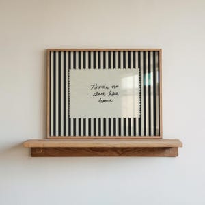 Puede incluir: Una impresión enmarcada con el texto "there is no place like home" sobre un fondo blanco, sobre un patrón de rayas blancas y negras. El marco es de color marrón claro y la impresión se muestra en una estantería de madera.