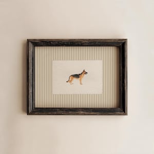 May include: A framed artwork featuring a watercolor painting of a German Shepherd dog. The dog is depicted in profile with black and tan fur. The frame is black with a distressed finish, and the background has vertical stripes.