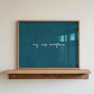 May include: A rectangular wall art piece featuring a deep teal background with the white cursive text "my cup overflows" centered in the middle. The artwork is enclosed in a light-toned wooden frame and rests on a matching wooden shelf.