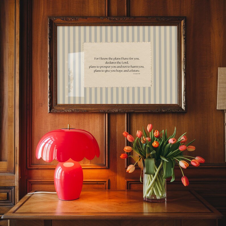 May include: A red mushroom-shaped table lamp with a scalloped edge sits on a wooden table. A framed print with vertical stripes and text is above the lamp. A vase of tulips is to the right.