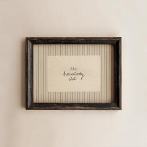 Puede incluir: Una obra de arte enmarcada con un marco de madera negro envejecido. La obra presenta un papel rectangular color crema con las palabras "the homebody club" en escritura negra. El fondo tiene rayas verticales.