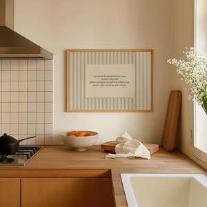 May include: A framed print with a light wood frame and a striped background. The print features a quote in a rectangular shape. A bowl of oranges, a cutting board, and a white cloth are on the wooden countertop.