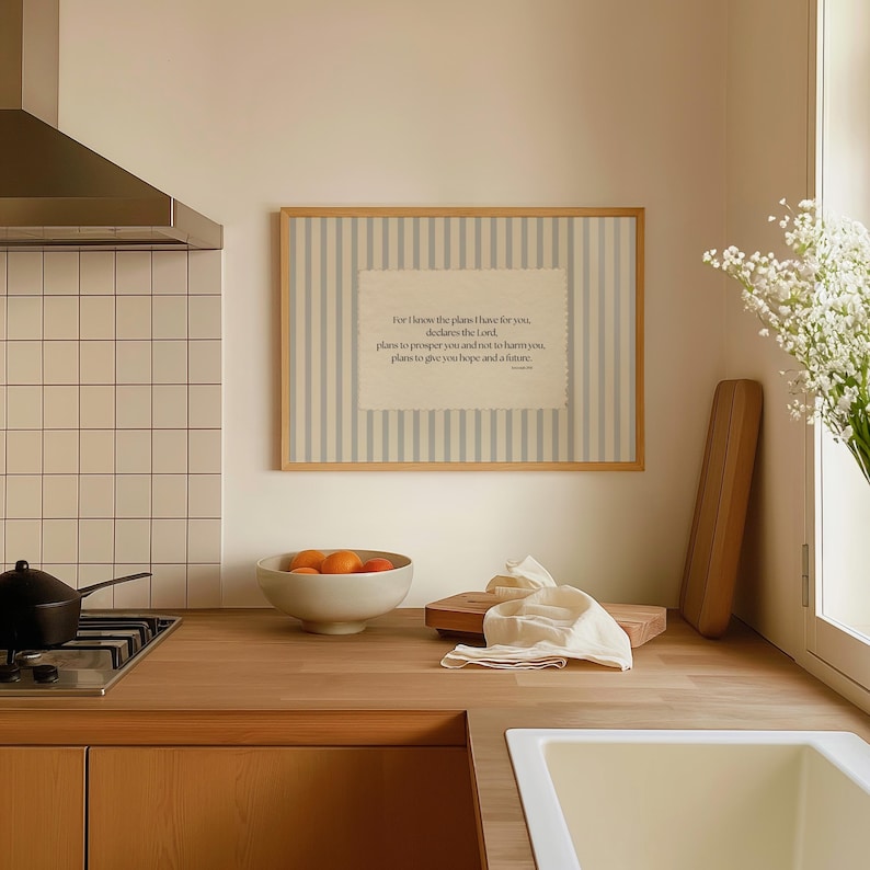 May include: A framed print with a light wood frame and a striped background. The print features a quote in a rectangular shape. A bowl of oranges, a cutting board, and a white cloth are on the wooden countertop.