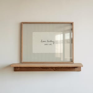 Puede incluir: Una obra de arte enmarcada con el texto "how lucky are we" en escritura negra. La obra de arte está sobre un fondo a rayas y enmarcada en un marco de madera marrón claro. Descansa sobre una estantería de madera a juego.