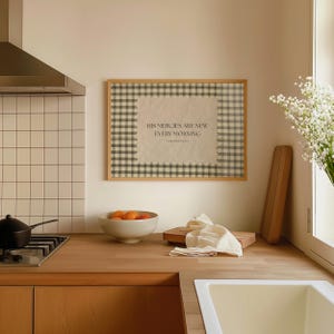 May include: A kitchen scene featuring a framed print with the text "HIS MERCIES ARE NEW EVERY MORNING". The print has a gingham border. A bowl of oranges, a wooden chopping board, and a white cloth are on the wooden countertop.