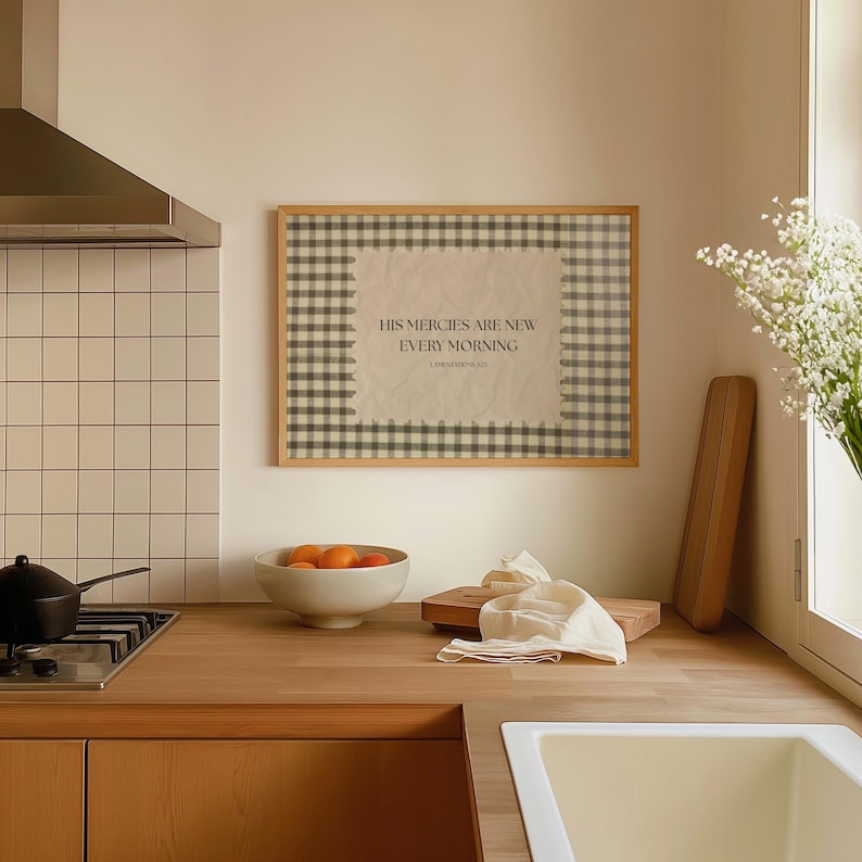 May include: A kitchen scene featuring a framed print with the text "HIS MERCIES ARE NEW EVERY MORNING". The print has a gingham border. A bowl of oranges, a wooden chopping board, and a white cloth are on the wooden countertop.