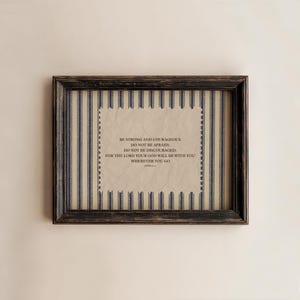 May include: A framed print with a black distressed wooden frame. The print has a cream-coloured background with blue and white vertical stripes. The text reads: "BE STRONG AND COURAGEOUS, DO NOT BE AFRAID; DO NOT BE DISCOURAGED, FOR THE LORD YOUR GOD WILL BE WITH YOU WHEREVER YOU GO."