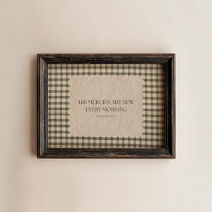 Puede incluir: Una impresión enmarcada con el texto "HIS MERCIES ARE NEW EVERY MORNING" en un marco negro desgastado. El fondo presenta un patrón de cuadros vichy en tonos beige y verde oliva.