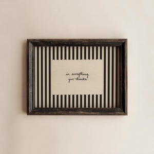 Puede incluir: Un marco rectangular de madera oscura desgastada que contiene una inserción de papel crema con el texto negro escrito a mano in everything give thanks, sobre un fondo de rayas verticales negras y blancas dentro del marco.