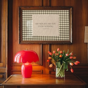 May include: A vibrant red mushroom-shaped lamp with a scalloped edge is on a wooden table. A framed artwork with a gingham pattern and the text "HIS MERCIES ARE NEW EVERY MORNING" hangs on the wood-panelled wall. A vase of tulips is also on the table.