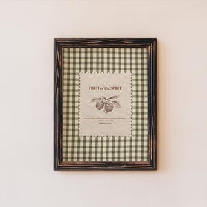 May include: A framed print featuring the text "FRUIT of the SPIRIT" with a botanical illustration of berries. The frame is dark brown with a distressed finish, and the background is a green and white gingham pattern.