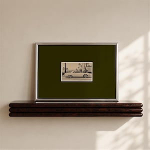 May include: A rectangular silver-toned frame containing a dark olive green mount and a centred black and white photograph of a vintage car parked on a street lined with palm trees. The frame is positioned horizontally with a clean, simple border.