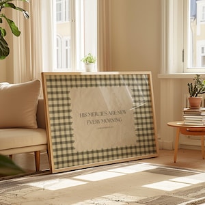 May include: A framed print with a light wood frame. The print features a gingham pattern in shades of grey and white, surrounding a central text that reads "HIS MERCIES ARE NEW EVERY MORNING". The artwork is displayed in a well-lit living room setting.