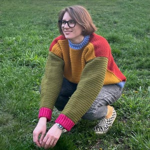May include: A colorful crochet sweater with a patchwork design. The sweater features a combination of red, yellow, green, and blue colors. The sleeves are green with pink cuffs, and the collar is blue. The person is wearing black-framed glasses and jeans.