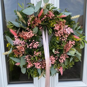 May include: A circular wreath featuring a mix of green eucalyptus leaves, dark green conifer sprigs, small yellow flowers, and clusters of pink baby's breath. The arrangement includes soft pink bunny tail grass and is suspended by a long, light pink ribbon.