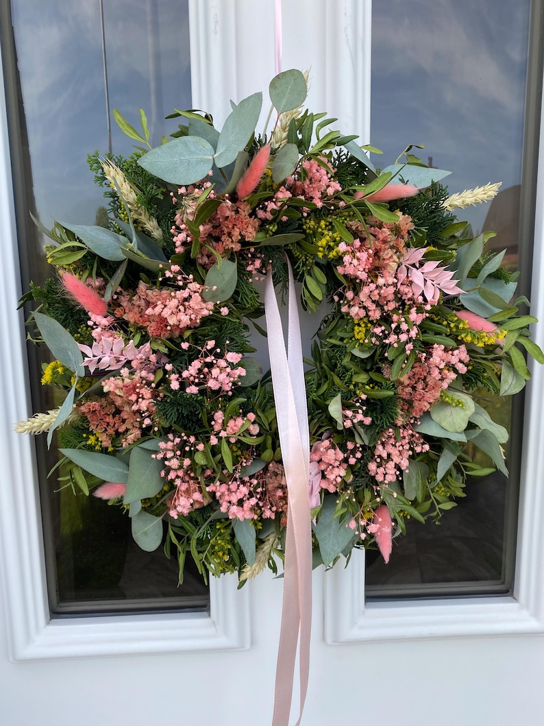 May include: A circular wreath featuring a mix of green eucalyptus leaves, dark green conifer sprigs, small yellow flowers, and clusters of pink baby's breath. The arrangement includes soft pink bunny tail grass and is suspended by a long, light pink ribbon.