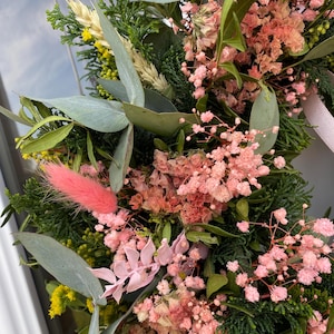 May include: A decorative wreath composed of dried flowers and greenery, featuring pink baby's breath, soft pink bunny tails, green eucalyptus leaves, dark green cedar sprigs, yellow solidago, and stalks of wheat, accented with a light pink ribbon.