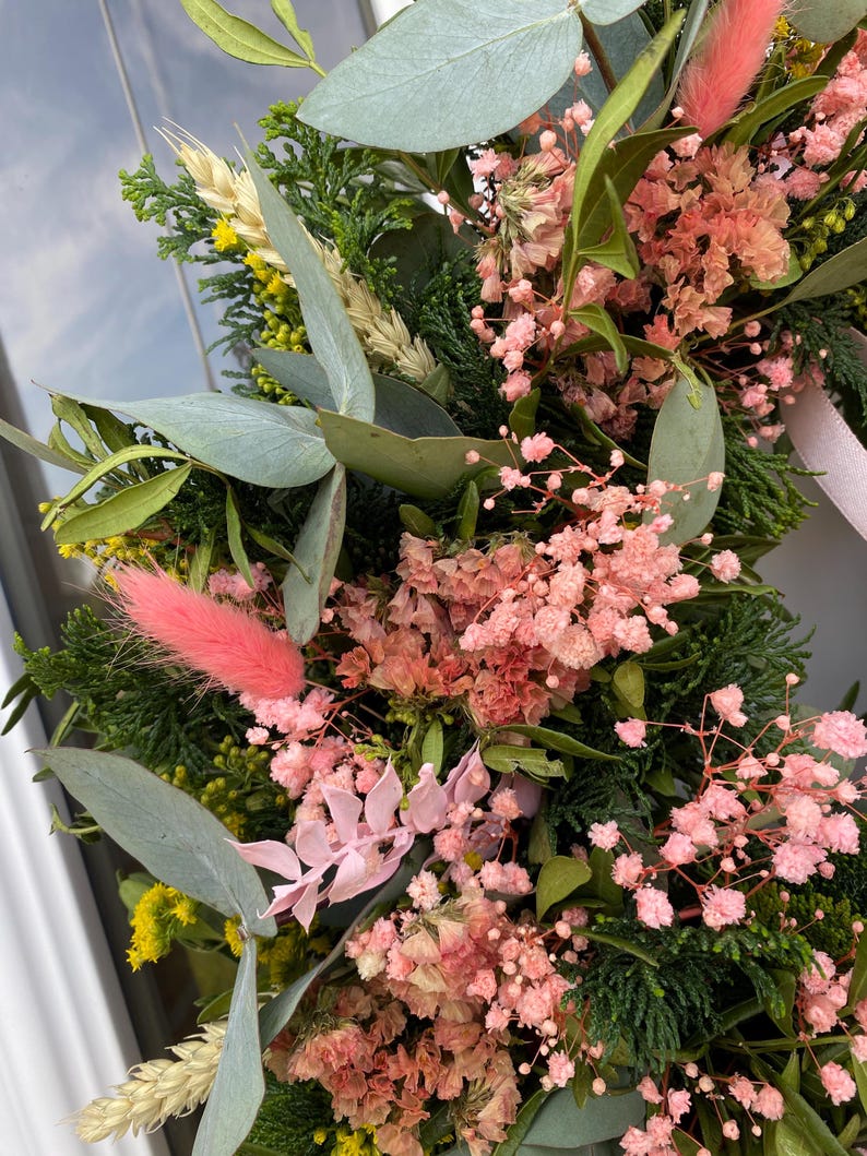 May include: A decorative wreath composed of dried flowers and greenery, featuring pink baby's breath, soft pink bunny tails, green eucalyptus leaves, dark green cedar sprigs, yellow solidago, and stalks of wheat, accented with a light pink ribbon.