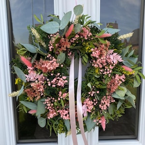 May include: A circular wreath composed of green foliage, eucalyptus leaves, and dried flowers in shades of pink and yellow. The arrangement features soft pink bunny tails and baby's breath, accented with two long, light pink satin ribbons hanging down.