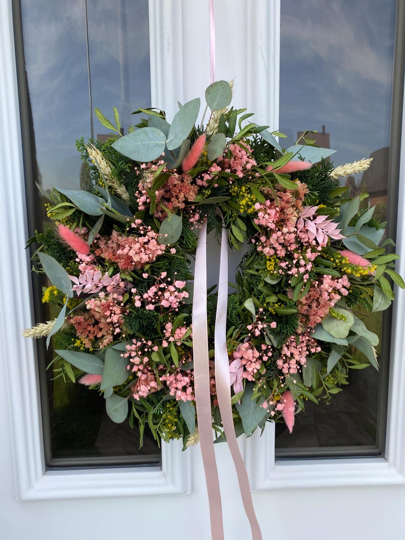 May include: A circular wreath composed of green foliage, eucalyptus leaves, and dried flowers in shades of pink and yellow. The arrangement features soft pink bunny tails and baby's breath, accented with two long, light pink satin ribbons hanging down.