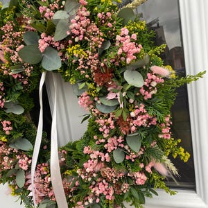 May include: A circular wreath constructed from green foliage, including eucalyptus leaves and cedar sprigs, accented with clusters of small pink baby's breath flowers, yellow filler flowers, and soft pink bunny tail grass, finished with a long white ribbon.