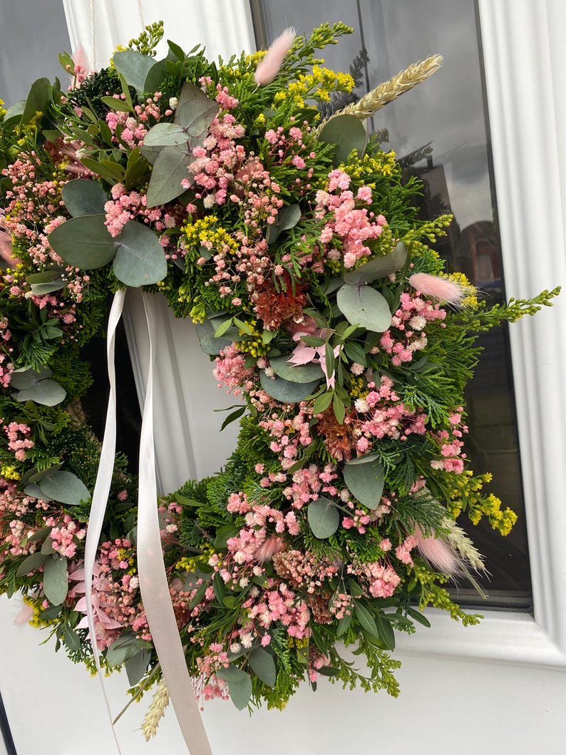May include: A circular wreath constructed from green foliage, including eucalyptus leaves and cedar sprigs, accented with clusters of small pink baby's breath flowers, yellow filler flowers, and soft pink bunny tail grass, finished with a long white ribbon.