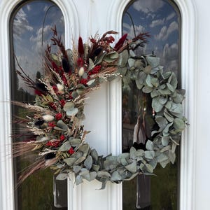Op de afbeelding: Een decoratieve krans gemaakt van gedroogde bloemen en bladeren. De krans bevat eucalyptusbladeren, rode bessen en diverse gedroogde grassen in bordeaux, zwart en cr&egrave;me tinten. De krans hangt aan een witte deur met boogvormige ramen.