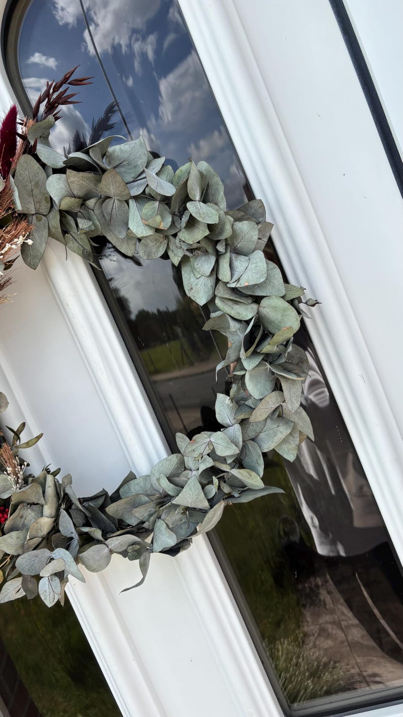 Op de afbeelding: Een cirkelvormige krans gemaakt van gedroogde eucalyptusbladeren en ander gedroogd blad. De krans heeft een gedempte groene kleur en wordt getoond op een witte deur met een glaspaneel. Een decoratief item.