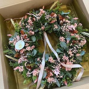 May include: A circular wreath composed of green foliage, pink baby's breath, dried wheat stalks, and small pink bunny tail grasses. The arrangement features a round white tag with a gold logo and two long, pale pink satin ribbons hanging down.