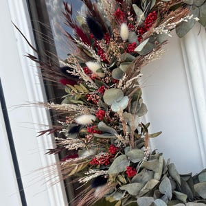 Op de afbeelding: Een droogbloemenkrans met een verscheidenheid aan texturen en kleuren. De krans bevat bordeauxrode, zwarte en witte accenten, met rode bessen en groene eucalyptusbladeren. De krans wordt op een witte deur getoond.