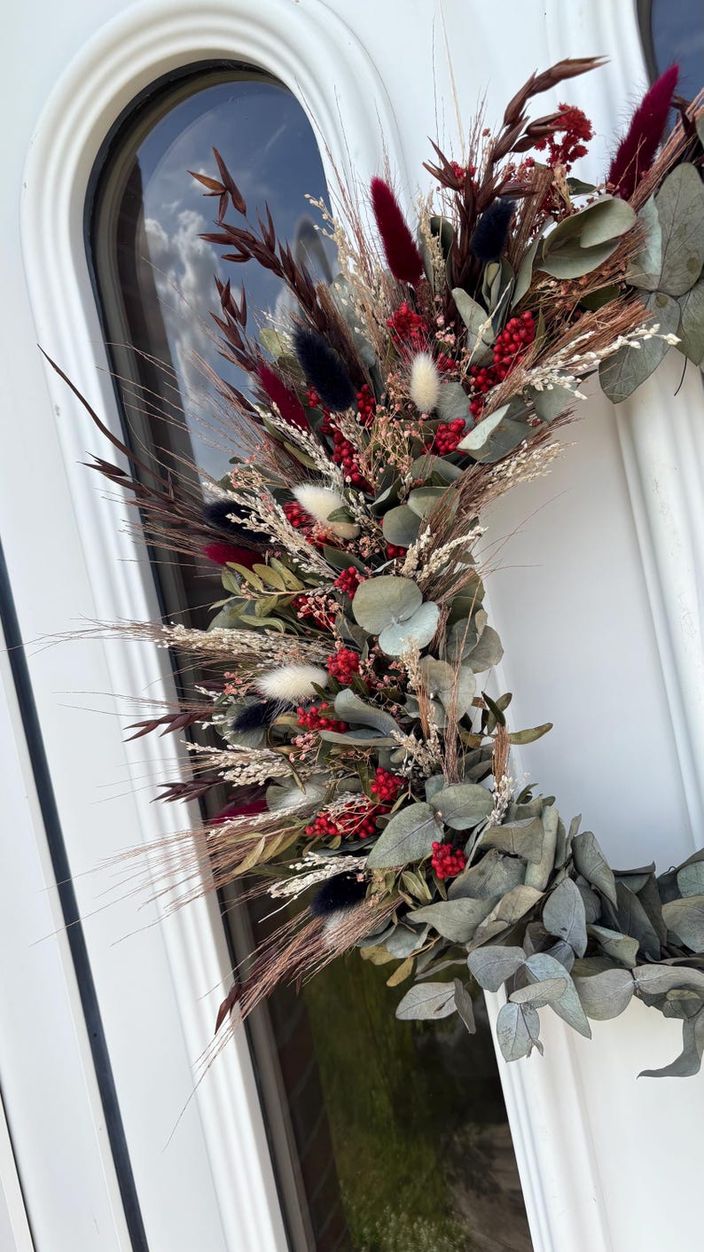 Op de afbeelding: Een droogbloemenkrans met een verscheidenheid aan texturen en kleuren. De krans bevat bordeauxrode, zwarte en witte accenten, met rode bessen en groene eucalyptusbladeren. De krans wordt op een witte deur getoond.