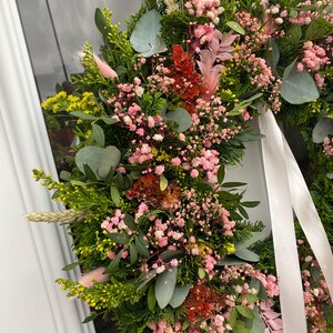 May include: A circular wreath composed of lush green foliage, including cedar and eucalyptus leaves, accented with clusters of pink baby's breath, yellow solidago, dried wheat stalks, and soft pink bunny tails, finished with a long white ribbon.