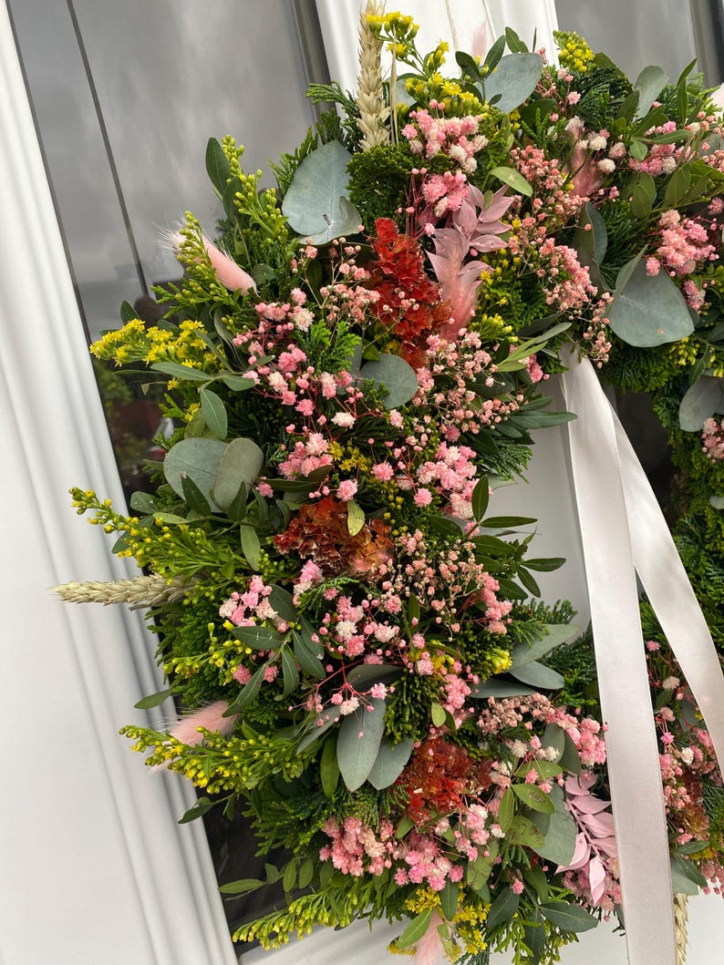 May include: A circular wreath composed of lush green foliage, including cedar and eucalyptus leaves, accented with clusters of pink baby's breath, yellow solidago, dried wheat stalks, and soft pink bunny tails, finished with a long white ribbon.