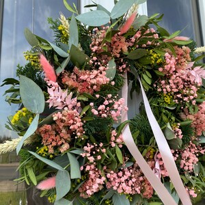 May include: A circular wreath featuring a mix of green eucalyptus leaves, dark green cedar sprigs, yellow wildflowers, and dried pink baby's breath. The arrangement includes soft pink bunny tail grass and is accented with long, light pink satin ribbons.