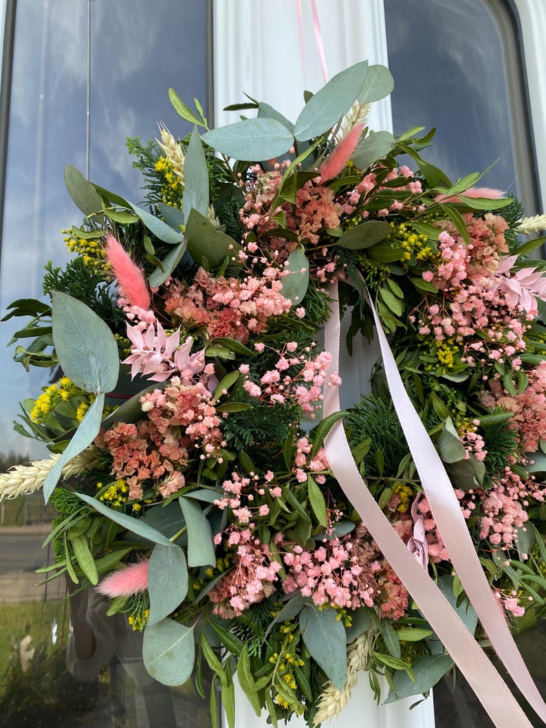 May include: A circular wreath featuring a mix of green eucalyptus leaves, dark green cedar sprigs, yellow wildflowers, and dried pink baby's breath. The arrangement includes soft pink bunny tail grass and is accented with long, light pink satin ribbons.