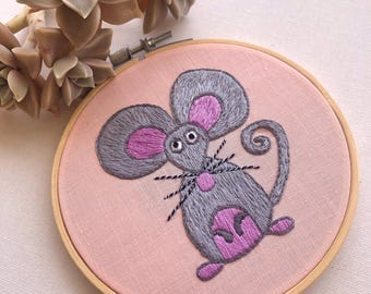 May include: Hand-stitched embroidery art featuring a gray mouse with pink accents on a pink fabric background. The mouse has large ears, a curly tail, and black whiskers. The embroidery is set in a wooden hoop, with a succulent plant in the background.
