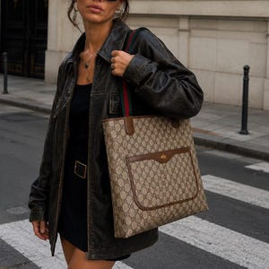 Puede incluir: Un bolso tote con estampado de monograma en beige y marrón, ribetes de cuero marrón, un bolsillo frontal y correas de hombro a rayas verdes y rojas. El bolso presenta un patrón repetitivo y un emblema dorado central en la solapa del bolsillo.