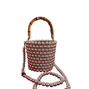 Puede incluir: Un bolso de mano de cuentas plateadas con asa de bambú y una larga correa de cuentas plateadas. La correa tiene un acento de cuentas marrones.