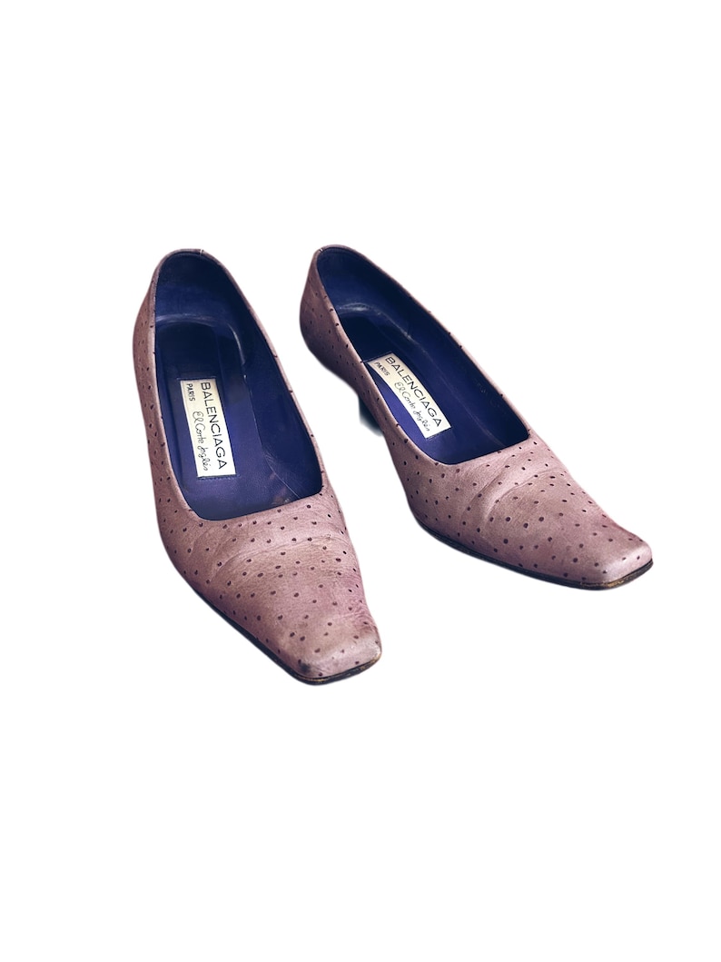 May include: A pair of light brown, square-toe pumps with small, dark dots. The shoes have a low heel and a purple interior. The brand name "BALENCIAGA PARIS El Corte Ingles" is printed inside each shoe.