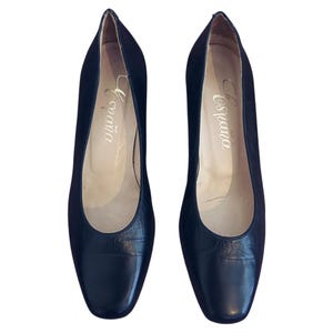 Puede incluir: Un par de zapatos de cuero negro con tacón bajo. Los zapatos tienen una punta redondeada y un tacón pequeño. La palabra "España" está impresa en dorado en la plantilla de cada zapato.