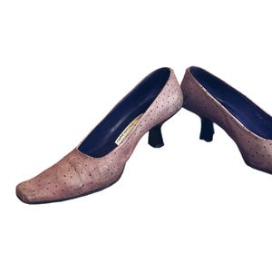 May include: A pair of light brown, heeled pumps with a square toe. The shoes have a textured surface with small, dark spots and a dark blue interior. The heels are black and the shoes appear to be made of leather.