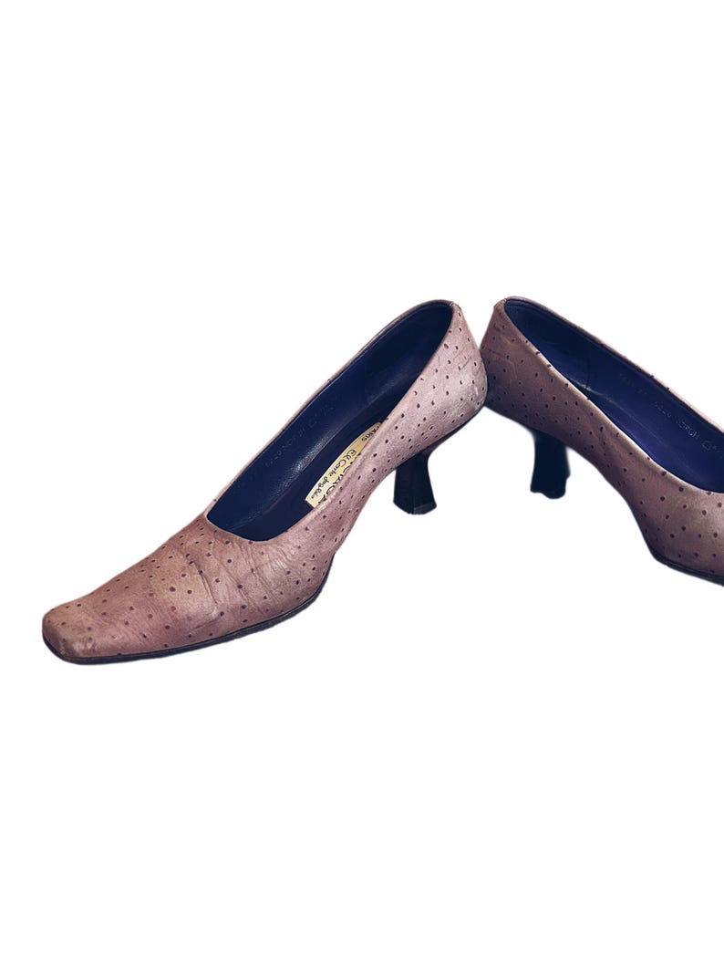 May include: A pair of light brown, heeled pumps with a square toe. The shoes have a textured surface with small, dark spots and a dark blue interior. The heels are black and the shoes appear to be made of leather.
