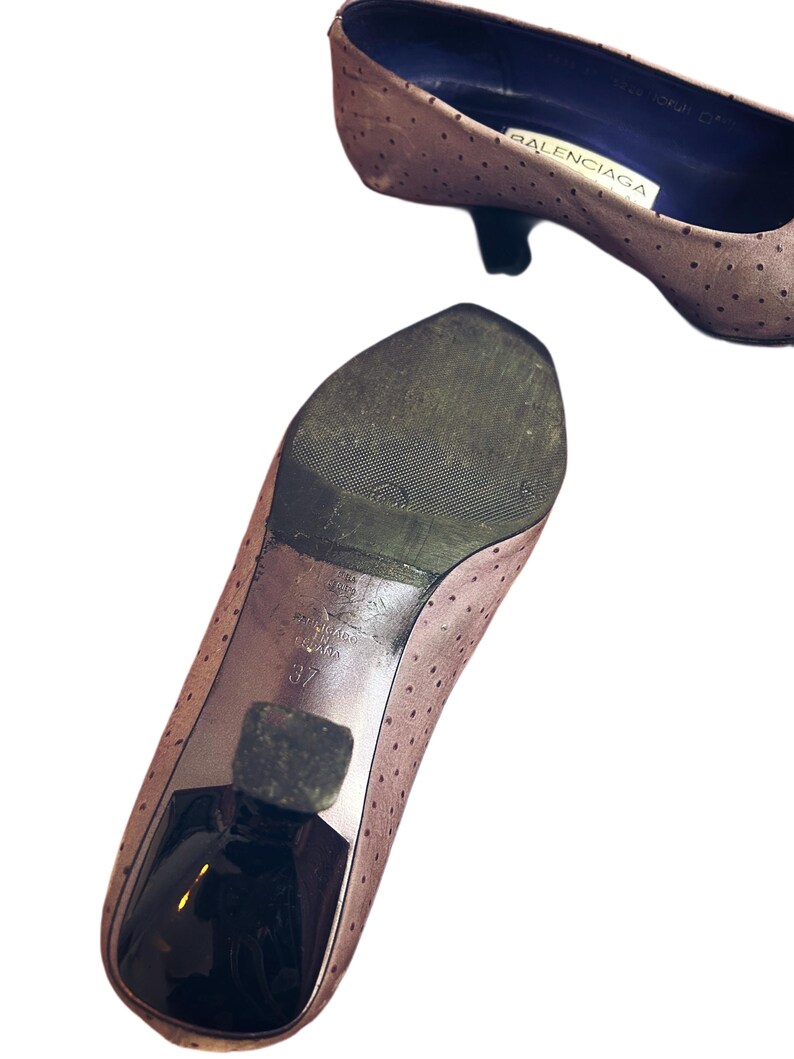 May include: Two light brown Balenciaga pumps with small black heels. The shoes have a perforated pattern and a pointed toe. The sole of one shoe is visible, showing the size 37 and the Balenciaga brand.